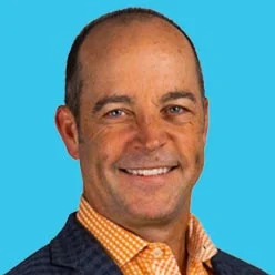 Professional headshot: smiling man in a navy blazer and orange check shirt, against a bright blue background.