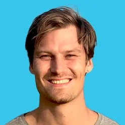 Close-up portrait of a smiling man with short brown hair against a bright blue background in a gray shirt.