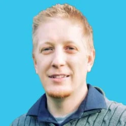 Portrait of a smiling man with blond hair wearing a green sweater over a collared shirt, against a bright blue background.