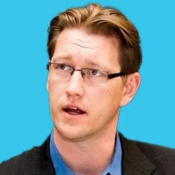 Man wearing glasses and a dark blazer speaks, with a solid bright blue background behind him.