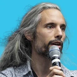 Man with long gray hair and stubble speaking into a microphone against a bright blue background, likely on stage.