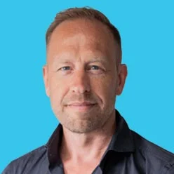 Professional headshot of a man with short hair and light stubble, wearing a dark collared shirt, against a bright blue background.